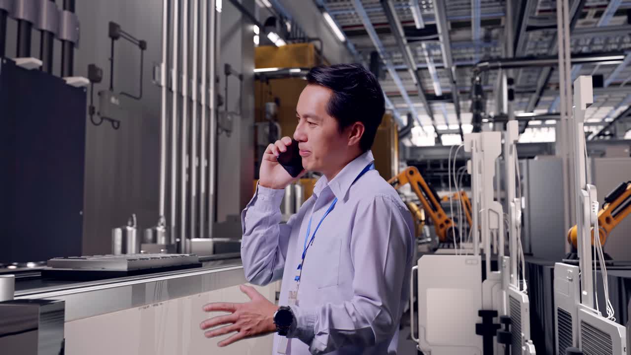 Vista lateral de un trabajador profesional asiático de pie, en una línea de producción automatizada de baterías en una fábrica moderna. Coge el teléfono y empieza a hablar con una ligera sonrisa.