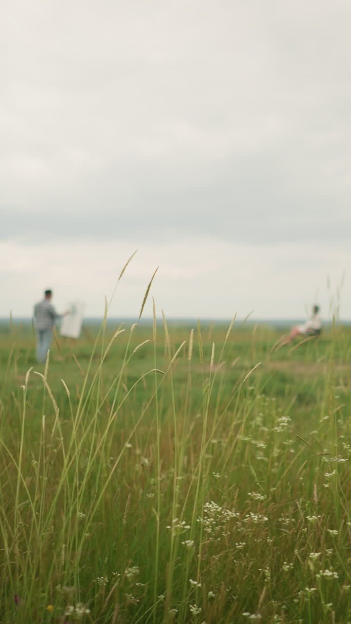 조용한 환경에서, 한 장인 (공예가) 이 높은 잔디 한가운데의 보드 위에 그림을 그리는 것을 볼 수 있습니다. 근처에, 한 여인이 의자에 편안하게 앉아 있습니다.