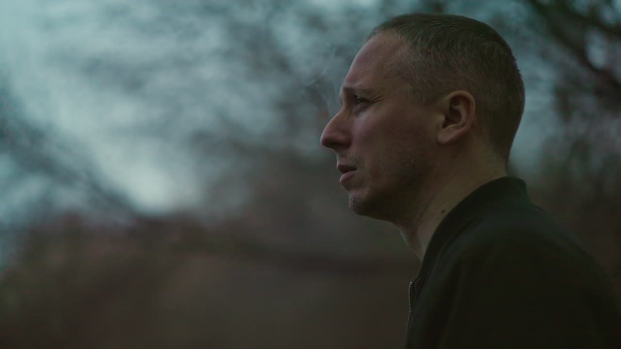 A man in a green jacket smokes a cigarette outdoors, with smoke visibly escaping his nose and mouth, the scene is shot at dusk, with a blurred background of trees