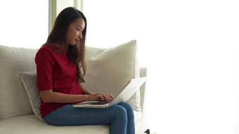 Trabajando-Desde-El-Concepto-De-Casa,-Mujer-Asiática-Sentada-En-Un-Sofá-Y-Escribiendo-En-Una-Laptop,-Toma-Estática-Con-Espacio-Para-Copiar