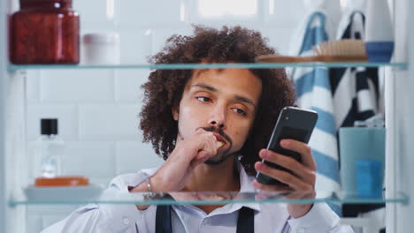 Blick-Durch-Den-Badezimmerschrank-Auf-Einen-Geschäftsmann,-Der-Zähne-Putzt-Und-Sein-Telefon-überprüft,-Bevor-Er-Zur-Arbeit-Geht