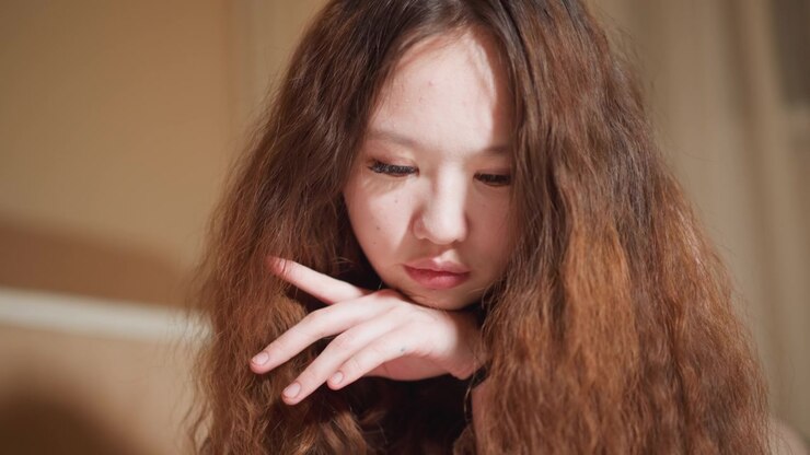 Close up portrait shows woman with loose waves in her brown hair. Her left hand rests gently against her chin and neck fingers slightly spread. background is softly blurred beige