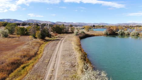 Vídeo-Aéreo-De-Un-Camino-Al-Lado-De-Un-Lago-Azul,-En-Un-Día-Soleado-Con-Montañas-En-La-Parte-Trasera