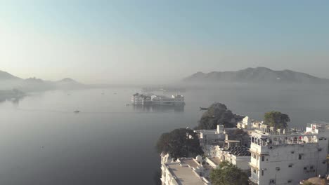 Taj-Lake-Palace-in-the-middle-of-the-lake-Pichola,-viewed-from-Ambrai-Ghat-in-Udaipur,-Rajasthan,-India---Aerial-slide-shot