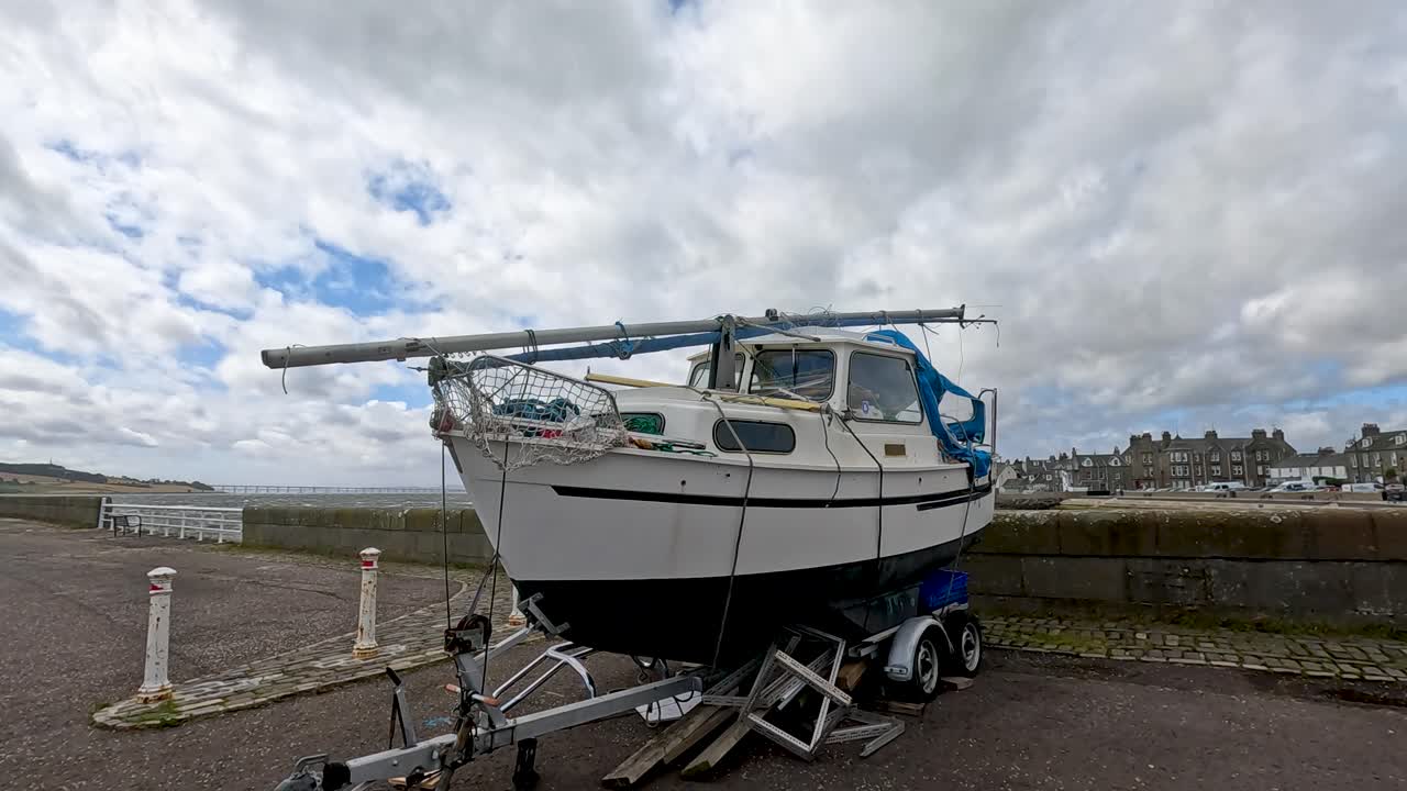 A small motor sailer boat on a trailer is stationary at a quayside under cloudy daylight, with the camera panning slightly for a wide, stable view