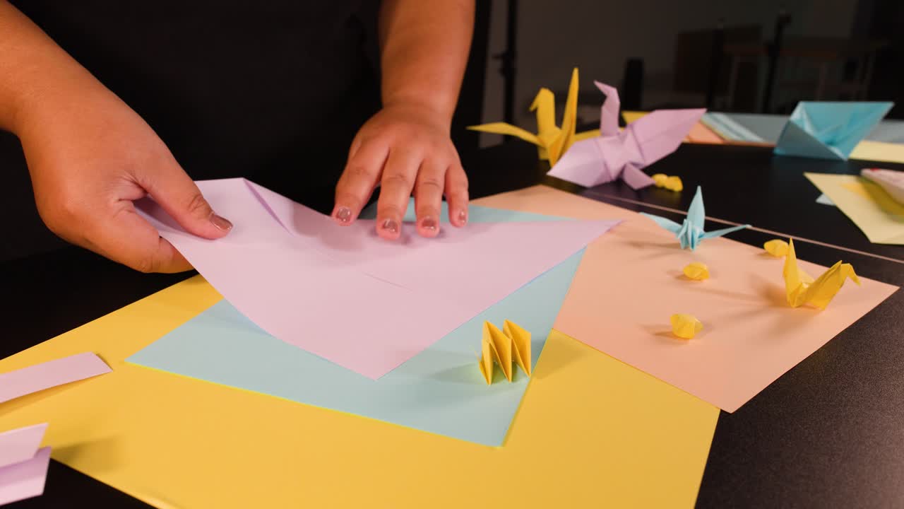 A person carefully folds pink paper on a desk surrounded by colorful origami paper cranes