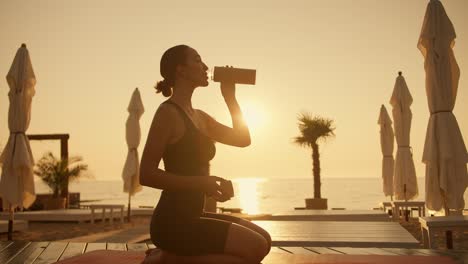 A-brunette-girl-in-a-black-sports-summer-suit-sits-on-her-knees-on-a-red-mat-and-drinks-water-from-a-sports-bottle-in-the-evening-during-a-golden-sunset-on-the-beach