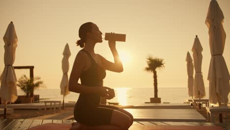 a brunette girl in a black sports summer suit sits on her knees on a red mat and drinks water from a sports bottle in the evening during a golden sunset on the beach