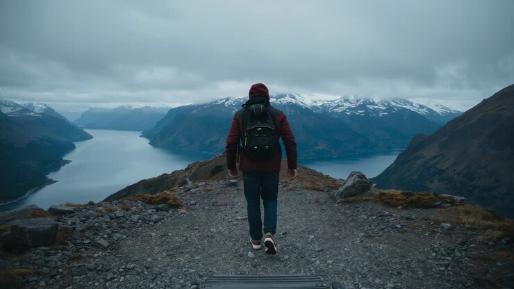 Walking hiker in maroon jacket prompted by trail heading to fjord viewpoint with black backpack