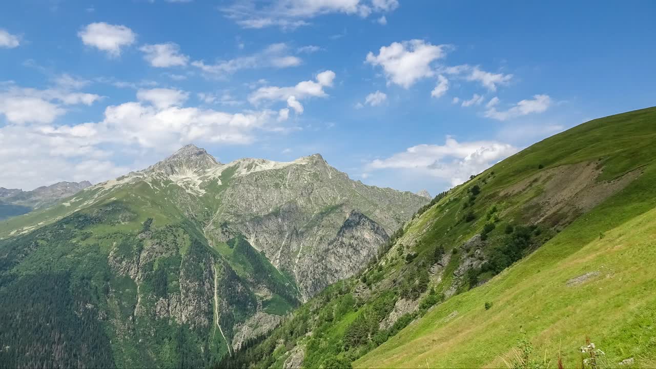 Rusya'da, dik yeşil yamaçlar, kayalık zirveler ve mavi gökyüzü altında hızla hareket eden bulutlarla bir Kafkas dağ vadisinin hızlandırılmış çekim görünümü, manzaralı doğa.