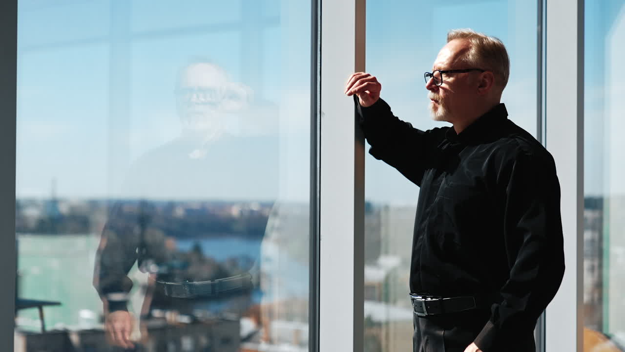 Serious adult man in black shirt and glasses walks along the panoramic window in the office. Thoughtful man stops and looks at the cityscape.