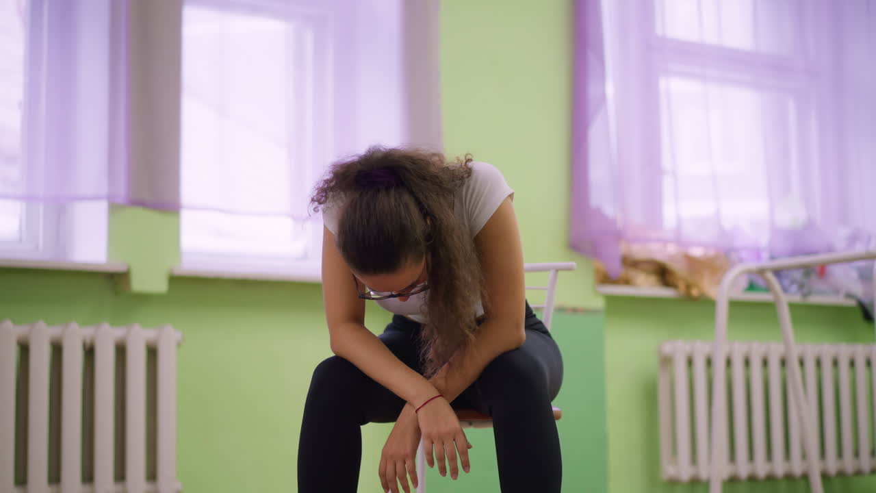 Young girl in white top and black leggings with sneakers sitting on chair, hands on knees, calm relaxed mood in studio with wooden floor, mirror reflection, soft green wall background after workout