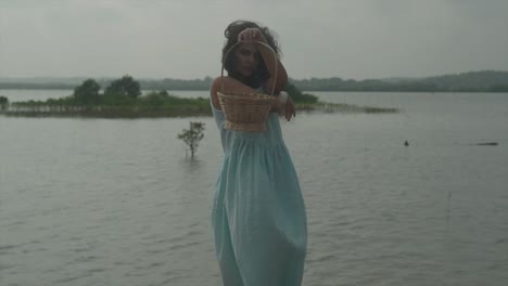 static slow motion shot of a young pretty woman in light blue dress and black hair standing in front of a lake with an island of plants and holding a wood basket in front of her