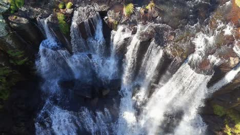 Drone camera descends above Ebor Falls, capturing cascading water, rocky cliffs, and lush vegetation in bright natural daylight with smooth downward movement