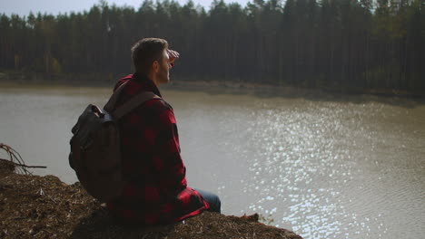 hombre con mochila viene se sienta en la orilla del lago hombre silueta pato salvaje cisne está nadando en el lago orilla cubierta de hierba verde reflejo del sol. imágenes de alta calidad 4k