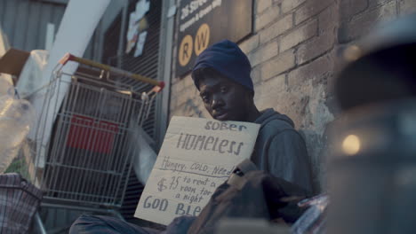 portrait of homeless black man holding handwritten sign in city alley