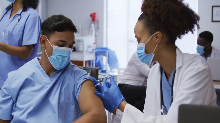 Mixed race female doctor giving covid vaccination to male colleague in hospital, both in face masks