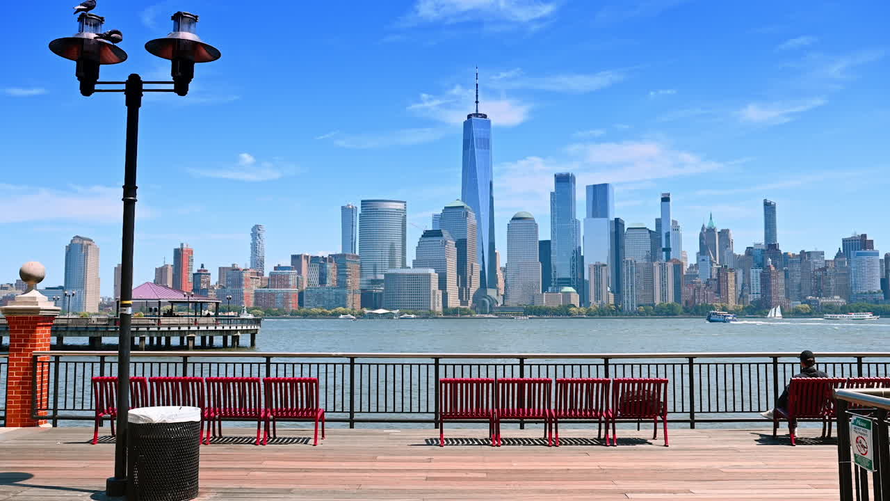 Walking up the railing and red benches on the pier along the Hudson River. View on the spectacular skyline of Manhattan on the other side of the river