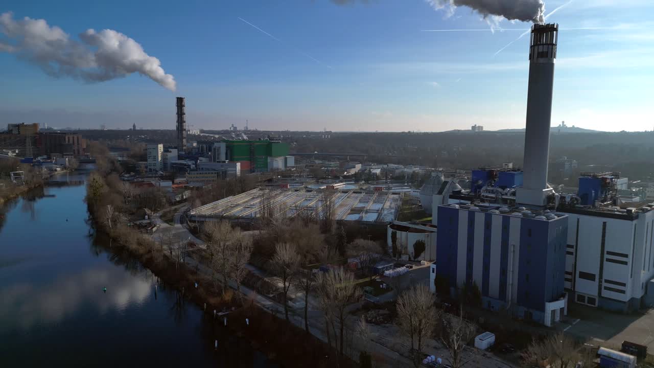 Waste to energy plant emitting smoke from its chimney, located near an urban area, environmental impact of waste. Smooth aerial view flight descending drone