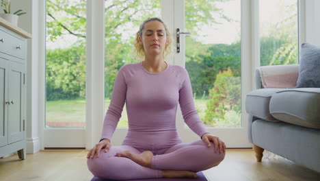 Portrait-Of-Smiling-Young-Woman-Sitting-On-Mat-At-Home-In-Yoga-Position-Meditating