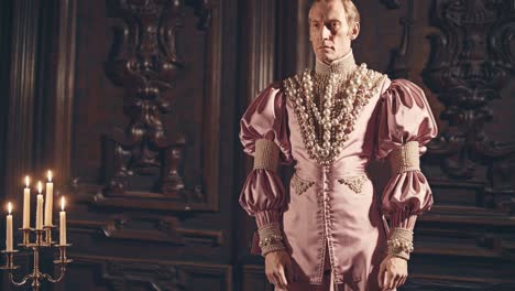 elegant nobleman posing in a dark vintage interior, wearing a pink renaissance costume with pearl necklaces and lit by candles on a candelabrum