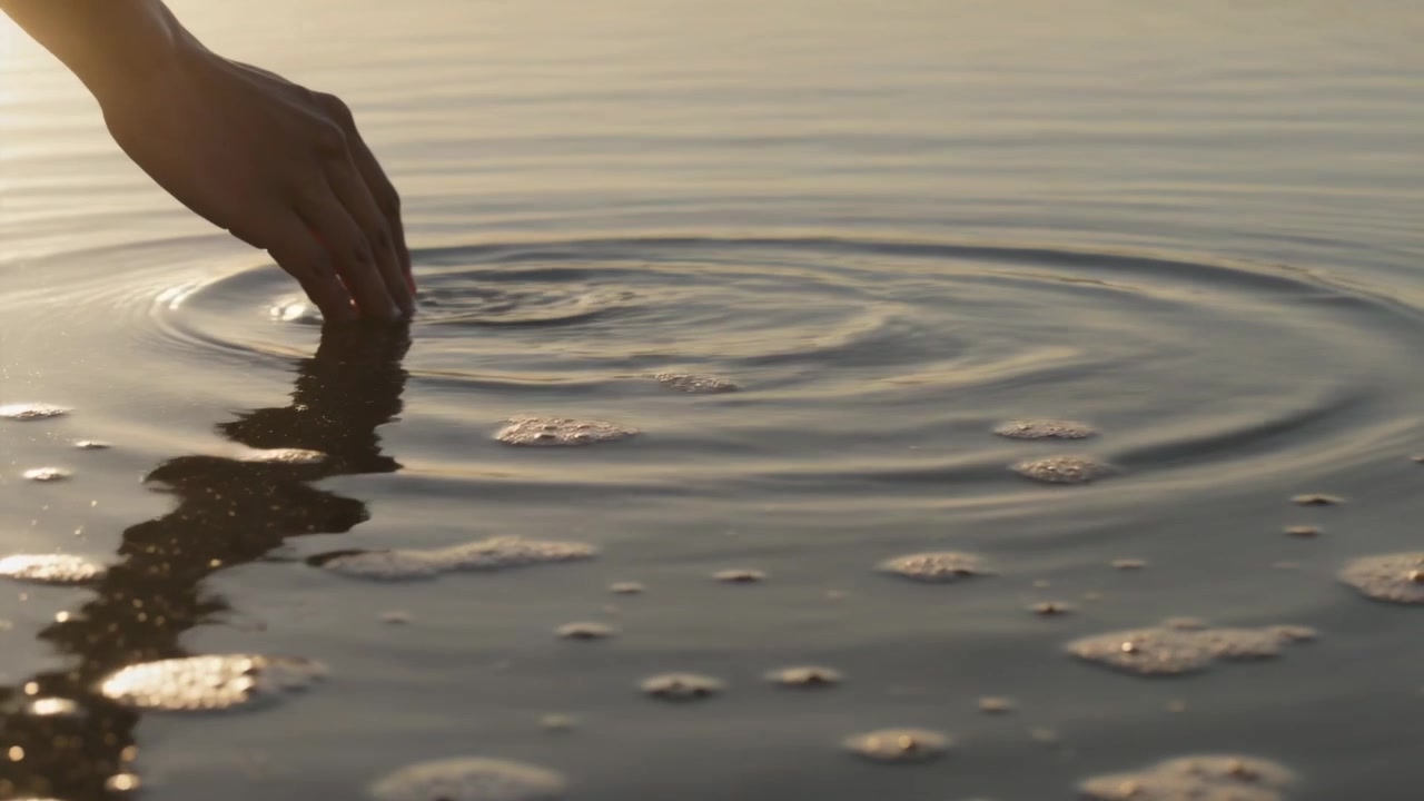 Lowering female hand touching shallow shoreline water on reflective sand, creating circular ripples