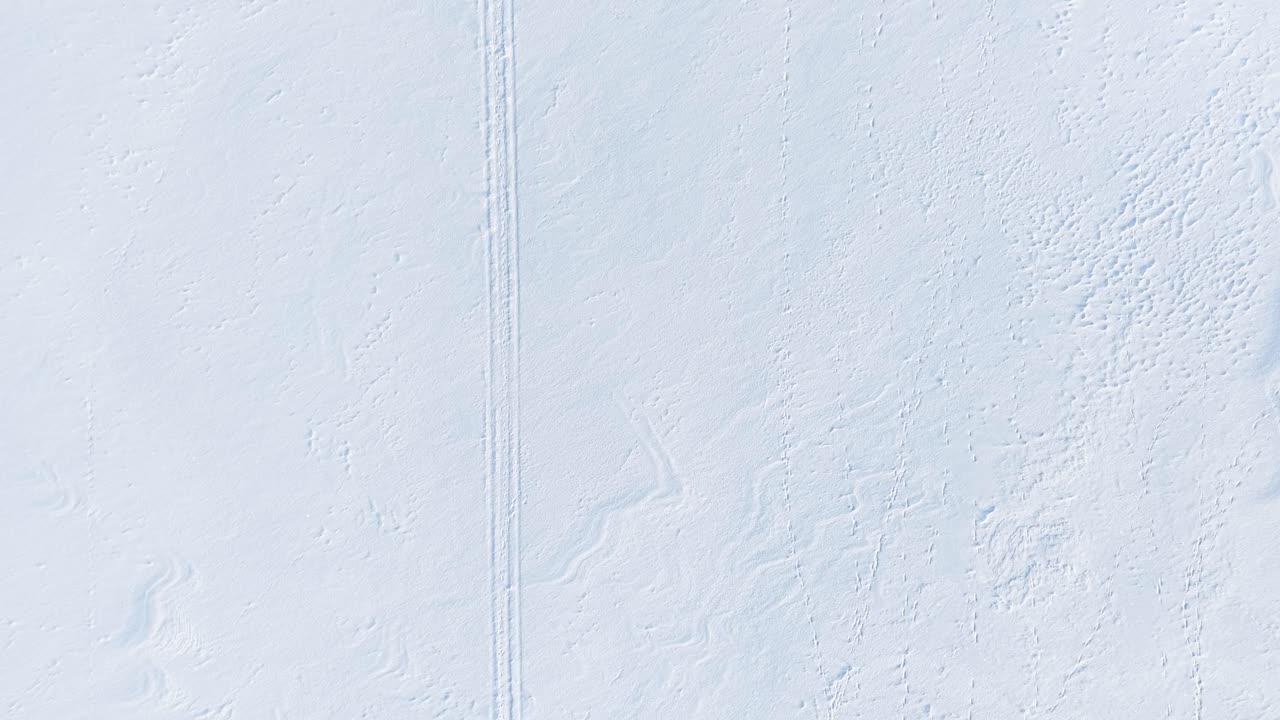 Erlebe eine atemberaubende Luftaufnahme einer weitläufigen, schneebedeckten Landschaft, die sich über ländliche Felder und eine kurvenreiche Straße unter sanftem Winterlicht erstreckt. Perfekt für eine ruhige Atmosphäre und natürliche Schönheit.