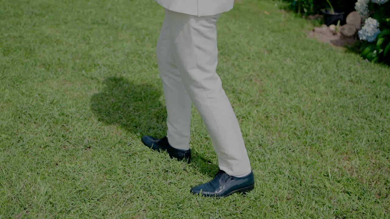 Groom in a white suit walking on grass, wearing dark blue leather shoes, with flowers in the background