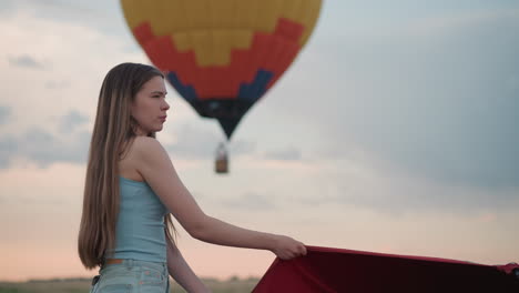 side view of young lady in denim shorts grips red hot air balloon panel as colorful envelope floats blurred in dusk sky during preflight beside rotating fan engine under pastel clouds