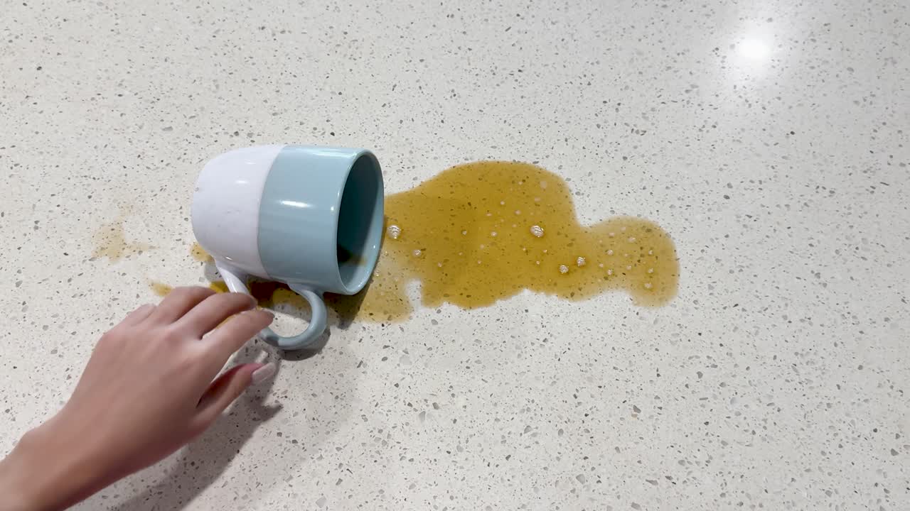 A hand accidentally spills coffee from a mug onto a kitchen countertop, creating a mess in bright lighting