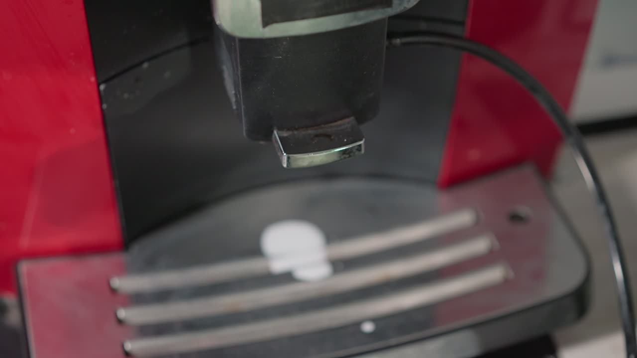 Close-up shot of espresso machine nozzle, focused on metal spout releasing a small droplet of coffee into a silver tray below. Modern kitchen appliance in action with shiny surface and chrome finish