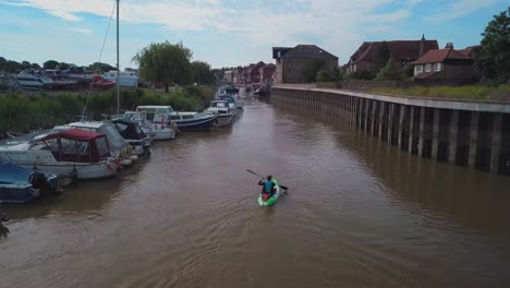 Man-kayaking-on-meandering-river-next-to-boats-and-houses,-drone-closely-follows-and-pans-up-to-reveal-beautiful-landscape