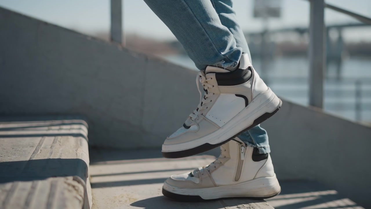 Kvindeben og fødder i blå jeans er fanget midt i et skridt på betontrapper, iført hvide høje sneakers med sorte detaljer. Baggrunden viser udendørs omgivelser med et vandområde.