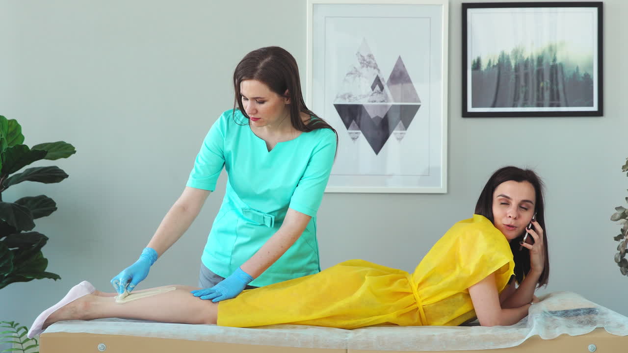 la chica habla por teléfono y sonríe durante la depilación de las piernas en el salón de belleza. depilación de las piernas con azúcar. maestro de depilación