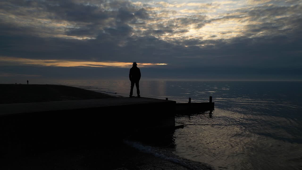 Uomo in silhouette in piedi sulla fine del molo al tramonto con piccole onde a Fleetwood Beach, Lancashire, Regno Unito.