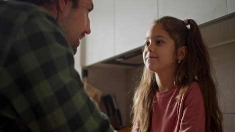 Close-up-shot-of-a-brunette-man-in-a-green-shirt-talking-to-his-little-brunette-daughter-in-a-pink-dress-who-is-sitting-on-the-table-in-the-kitchen-and-thinking-about-something