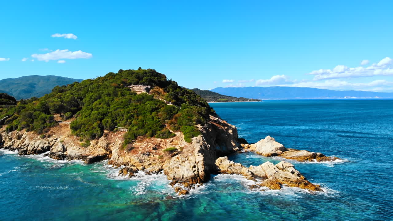 Aerial drone view of the Aegean sea rocky coast, peninsula with greenery and ancient excavations on the hill, waves with a lot of foam, Greece