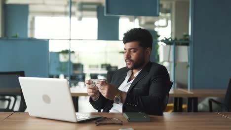 Retrato-De-Un-Joven-Apuesto-Y-Elegante-De-Apariencia-árabe-Sentado-En-Una-Oficina-Moderna-De-Un-Centro-De-Negocios,-Jugando-En-Su-Teléfono-Inteligente-Y-Celebrando-Su-Victoria