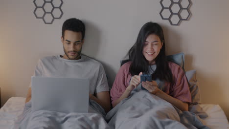 couple relaxing in bed, using laptop and smartphone