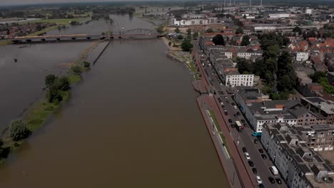 Descenso-Aéreo-Y-Panorámica-Lateral-Que-Muestra-El-Paisaje-Urbano-De-Zutphen,-Los-Países-Bajos,-Con-El-Tráfico-Que-Pasa-Por-El-Bulevar-Durante-Los-Altos-Niveles-De-Agua-Del-Río-Ijssel-Llenando-Las-Llanuras-Aluviales