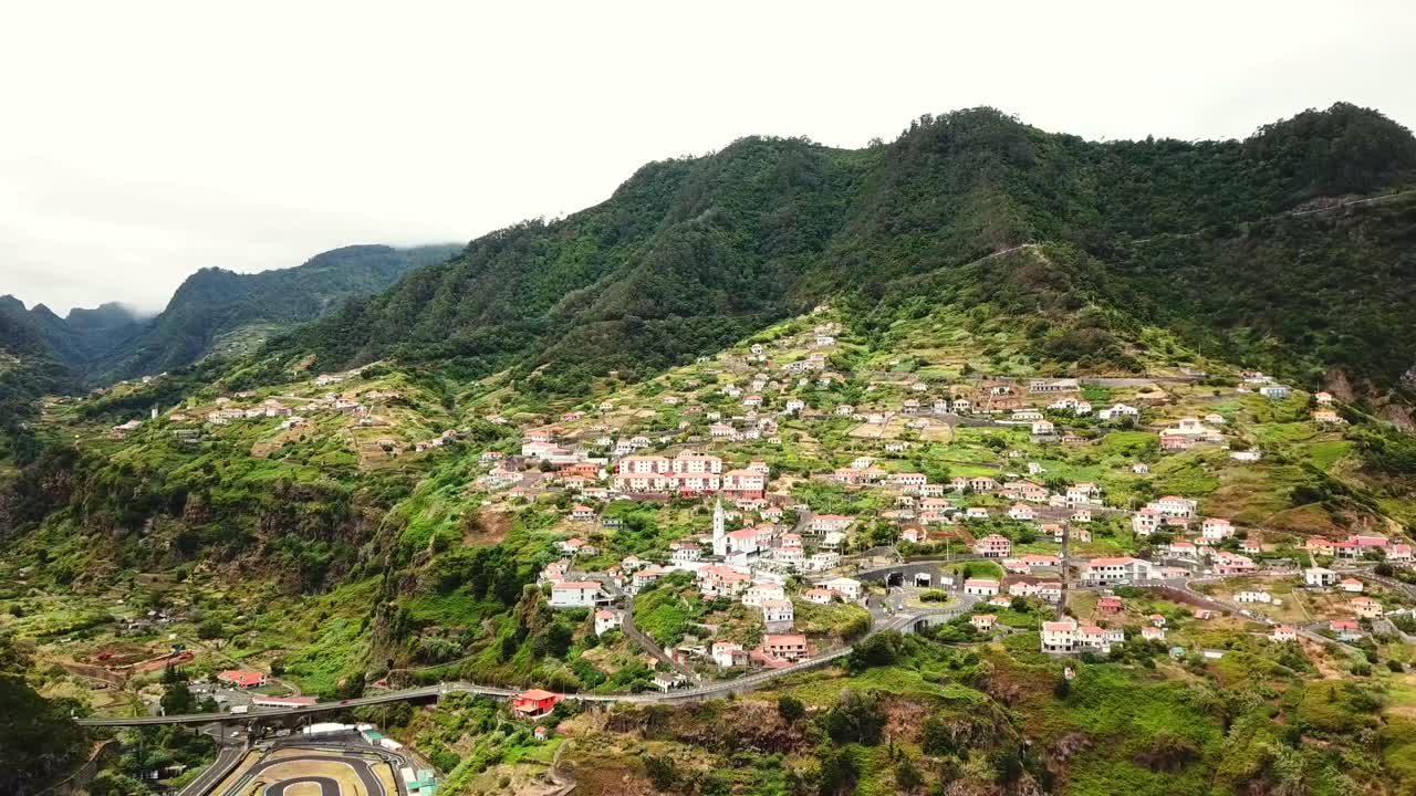 A breathtaking view of Faial, Madeira, nestled in a verdant valley surrounded by lush, forested mountains. The serene village features terraced slopes, charming houses, and winding roads