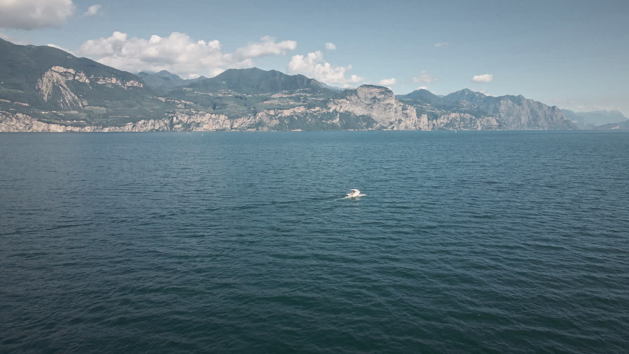 Volo drone 4K fluido in avanti sopra il Lago di Garda, che avanza verso una barca solitaria in acque aperte con creste montuose e scogliere drammatiche all'orizzonte sotto un cielo limpido