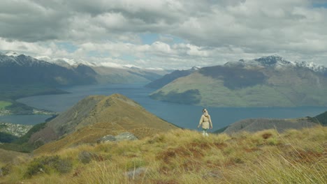 Woman-reaching-rock-outcrop-in-mountains-with-view-of-Queenstown-and-Lake-Wakatipu