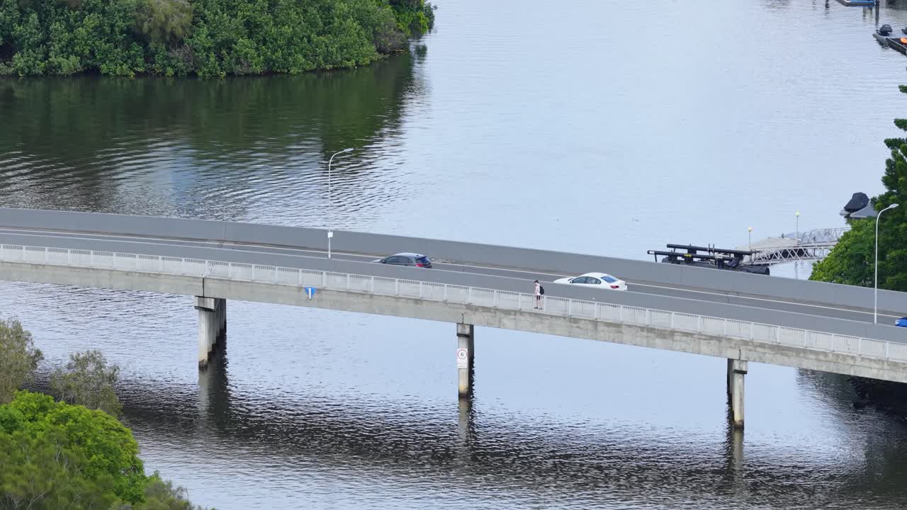 Cars travel across a bridge over a serene canal, surrounded by lush greenery, under clear daylight