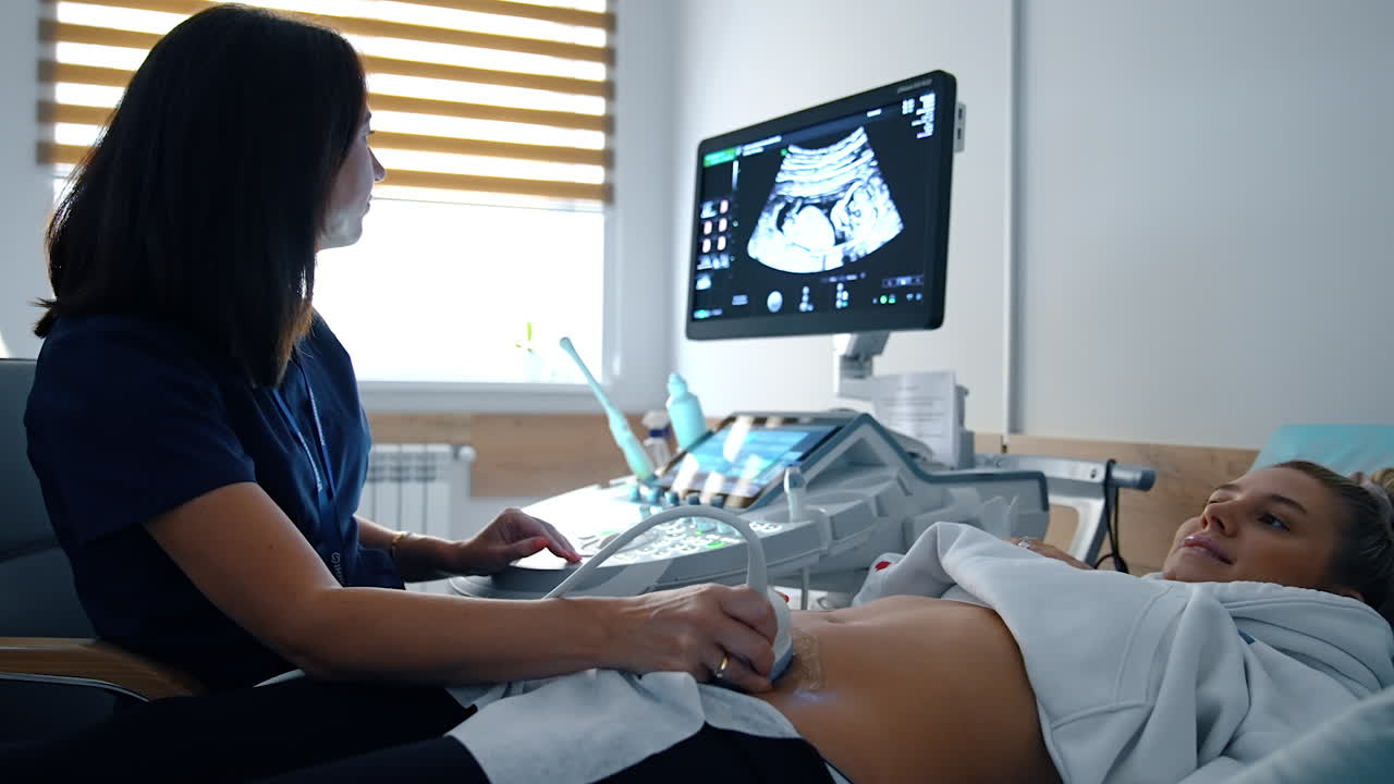 Female doctor performs ultrasound examination for a Caucasian woman. Pregnant lady smiles seeing her future baby on the screen.
