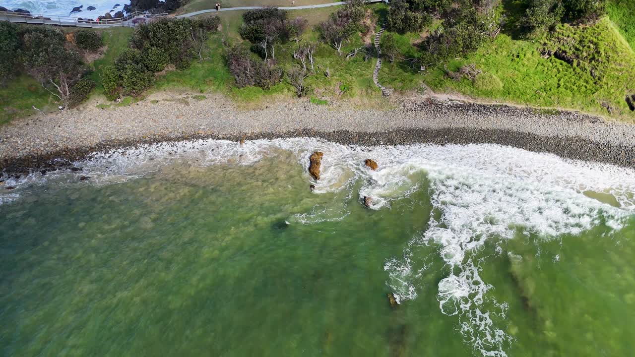 Drone captures sweeping views of Byron Bay's coastline, highlighting lush greenery, rocky shores, and crashing waves in vibrant daylight