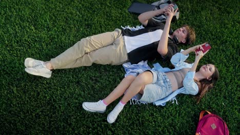 una pareja joven acostada en la hierba, sonriendo y mirando sus teléfonos.