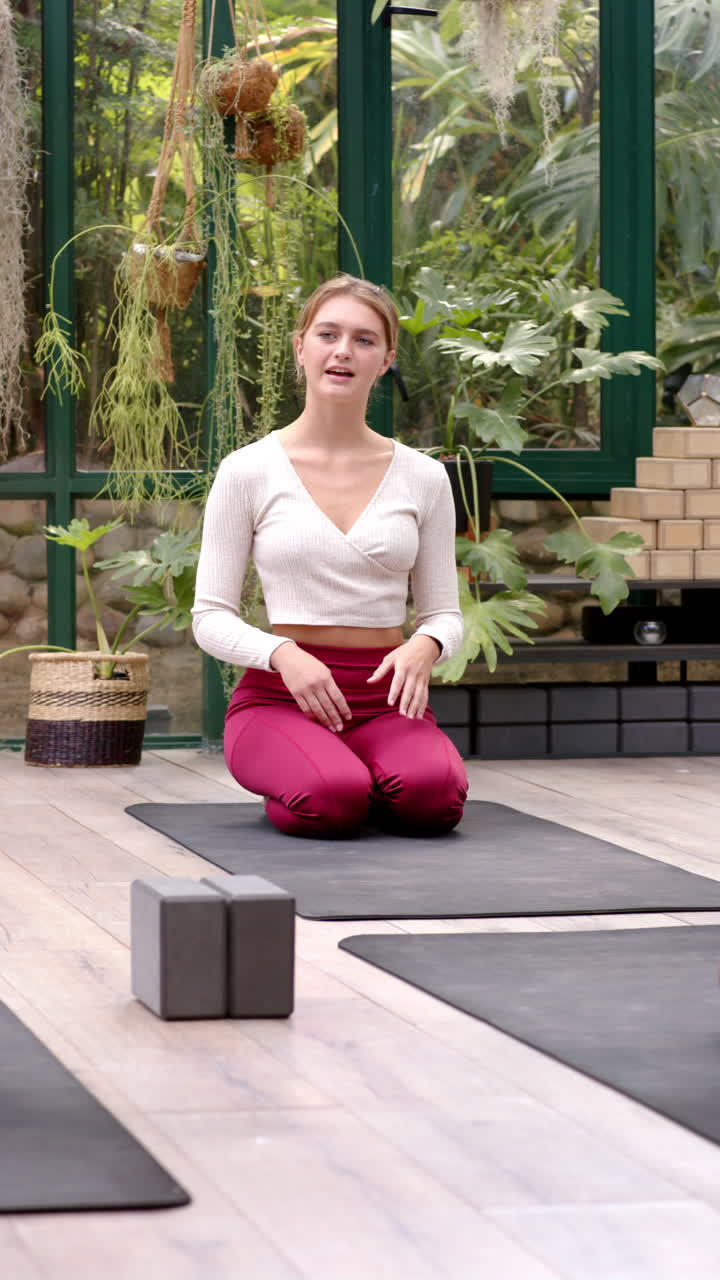 Vertical video: Practicing yoga, woman kneeling on mat in glass house, focusing on breathing