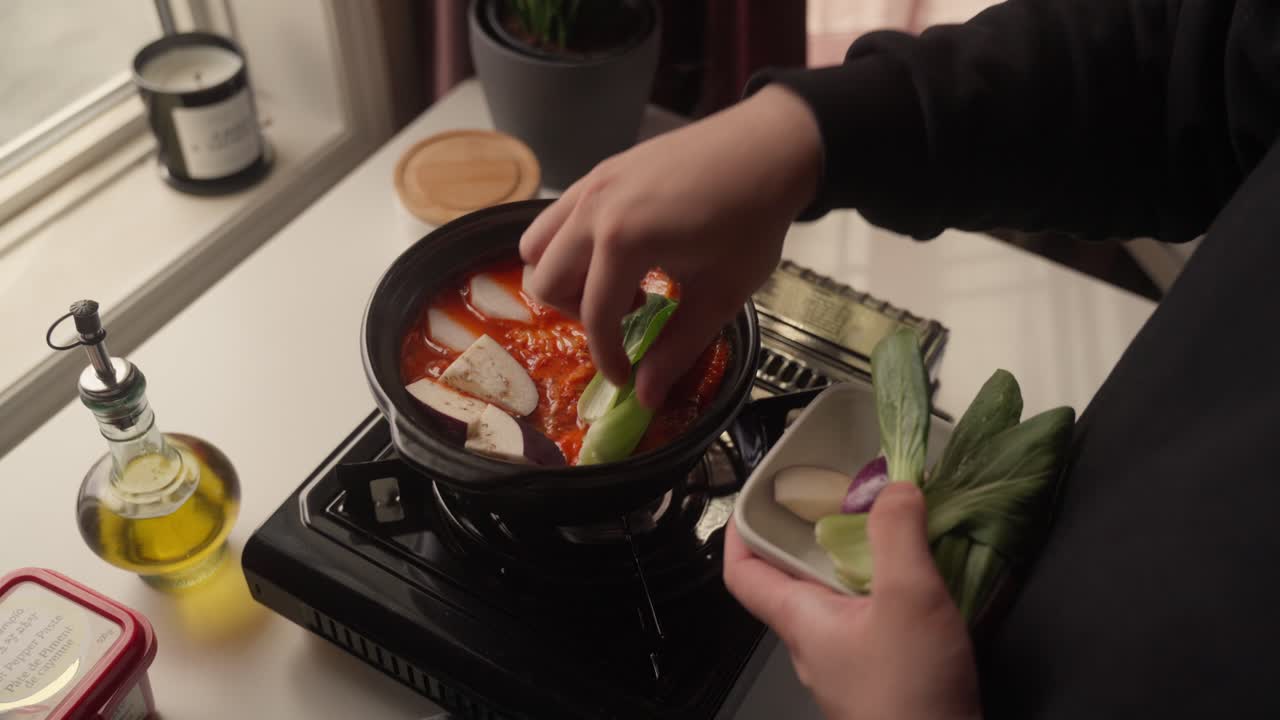 キッチン窓のそばで、ポータブルコンロで調理中の赤いスープ入り土鍋に野菜を入れる人物。温かい手作り料理を準備している。