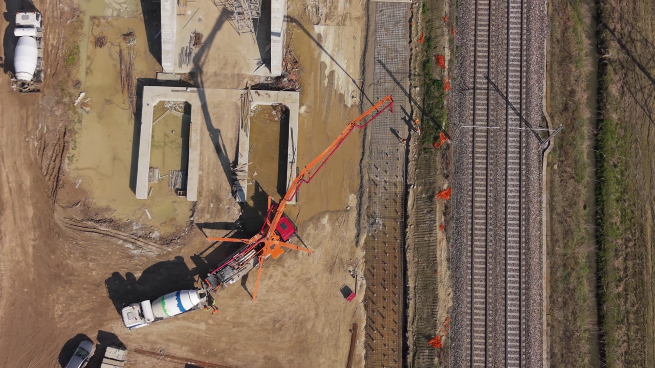Aerial drone view of a cement truck pouring concrete through a long pump pipe, delivering material across a new railway construction site in Italy.